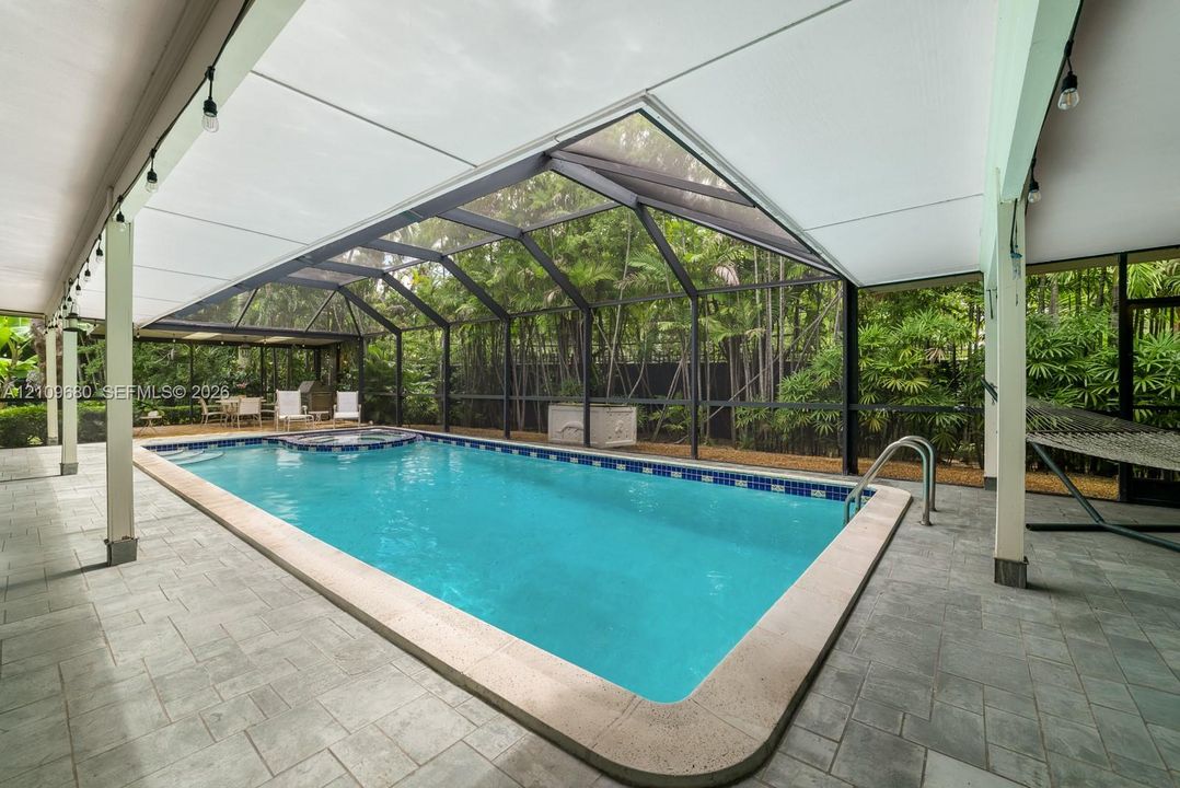 Screened-in pool area featuring an integrated spa, stone-tile decking, covered lanai, and surrounding lush greenery