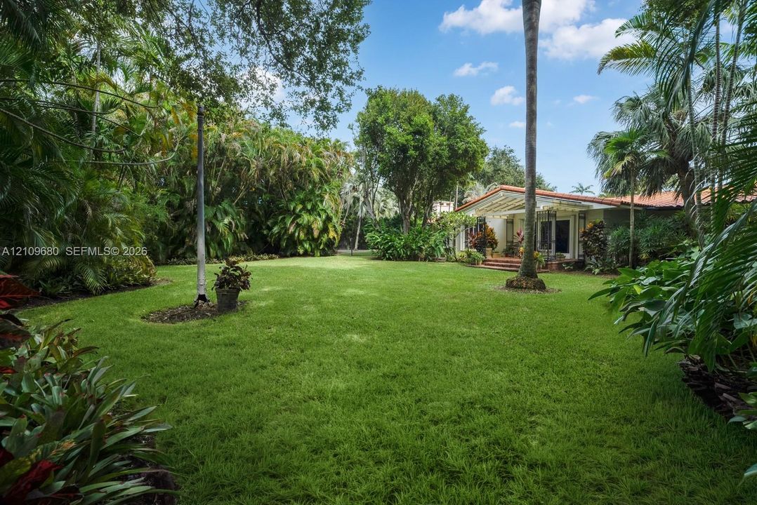 Expansive backyard featuring a manicured lawn, mature trees, dense tropical foliage, and a clay tile roof structure