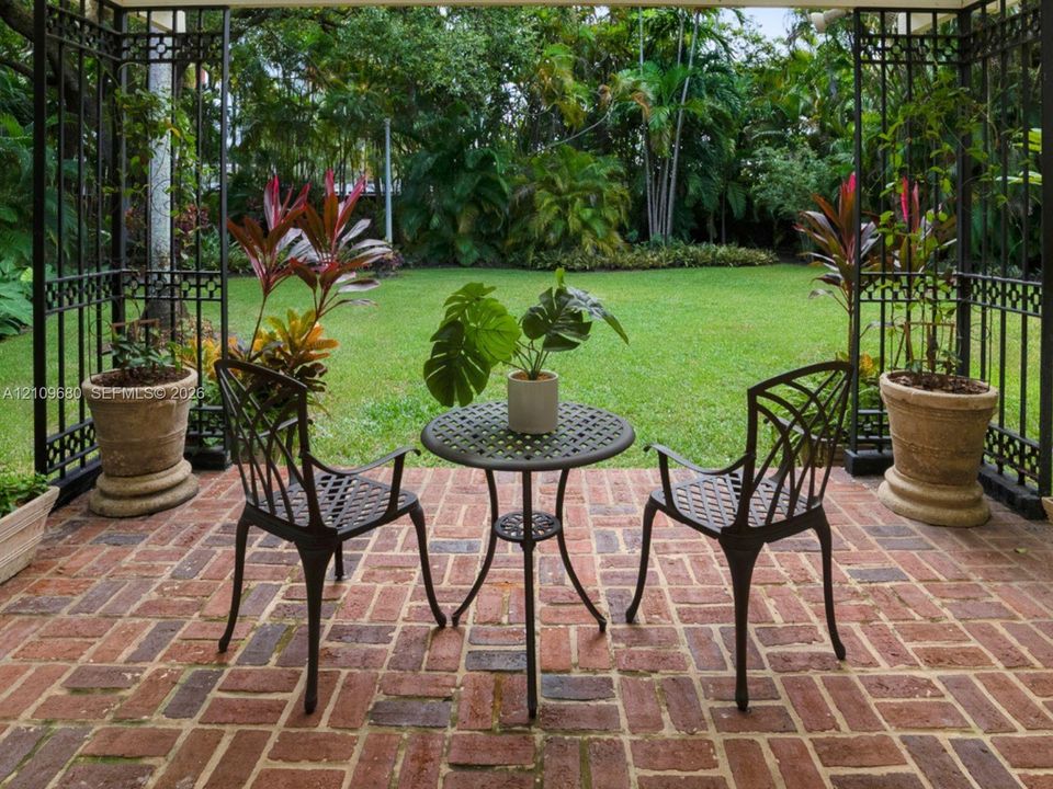 Covered patio featuring red brick paving, intricate black metal structural framing, and a level lawn bordered by mature landscaping