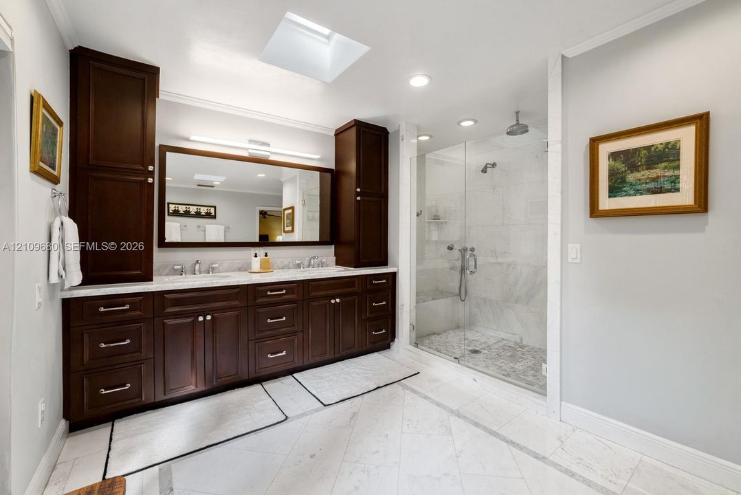 Bathroom featuring dark wood-finish cabinetry, dual undermount sinks, a walk-in shower with stone tile surround, and a ceiling skylight