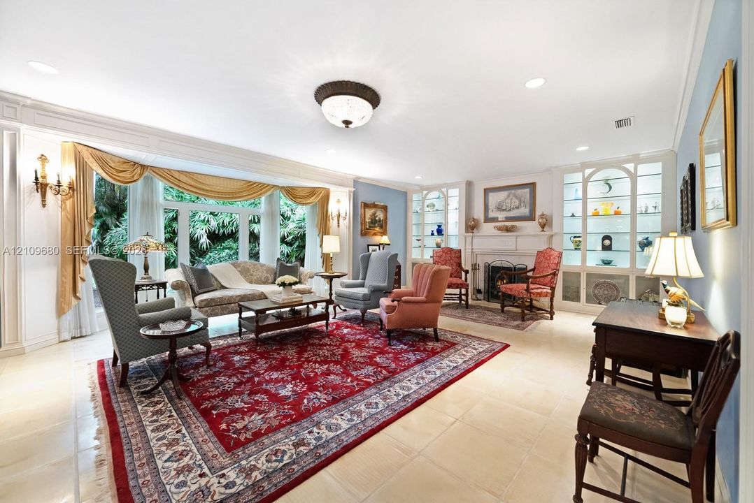 Spacious living area featuring large windows with detailed trim, built-in floor-to-ceiling glass display shelving, a fireplace with a decorative mantel, and light-toned tile flooring