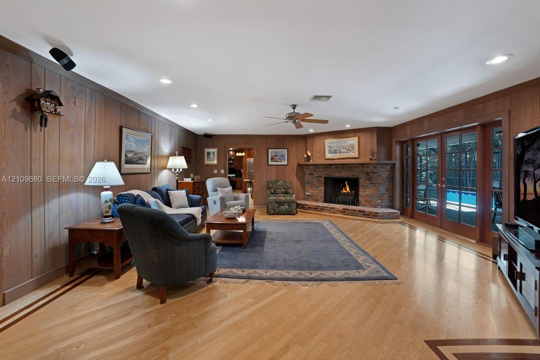 Living area featuring wood-paneled walls, wood flooring with inlaid border details, a corner stone fireplace with a raised brick hearth, a ceiling fan, and double glass-paneled doors