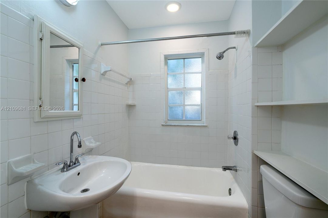 Bathroom featuring white wall tiling, a pedestal sink with a dual-handle faucet, a built-in bathtub with a shower head, integrated wall shelving, and a medicine cabinet