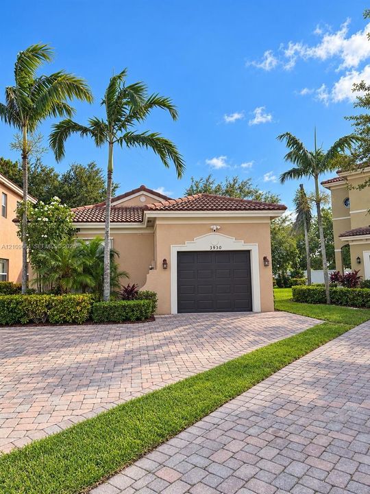 Exterior facade featuring a tile roof, stucco finish, single-car garage, and a paved stone driveway complemented by established palm trees and manicured hedges