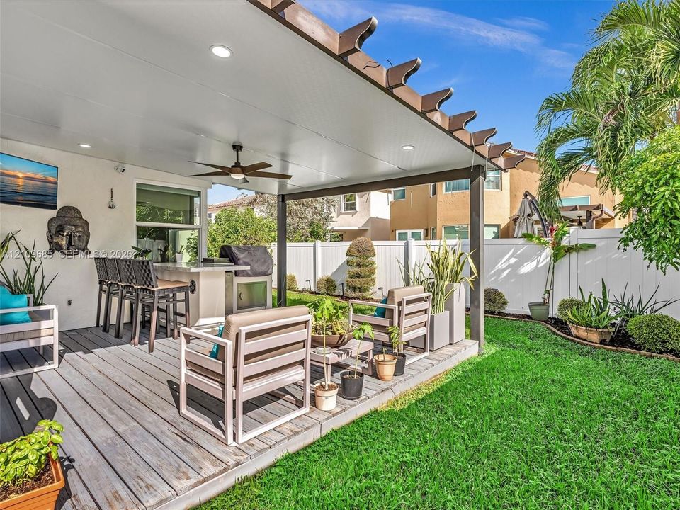 Covered outdoor patio featuring a wooden pergola, built-in stone grill station, recessed lighting, and a ceiling fan, opening onto a landscaped backyard with a privacy fence