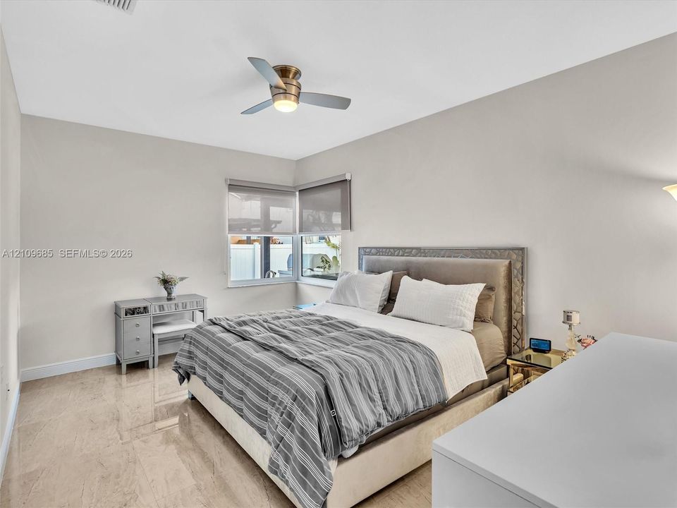 Polished stone flooring, neutral wall tones, multi-pane corner window configuration with roller shades, and a ceiling-mounted light fixture with a three-blade fan
