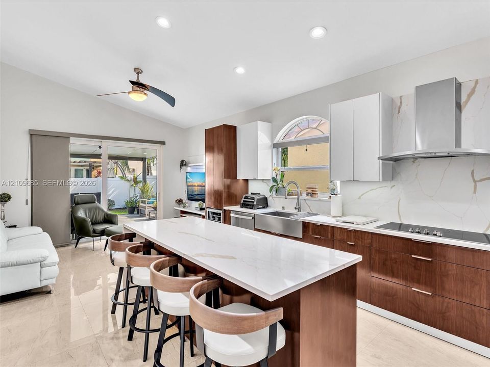 Kitchen featuring two-tone flat-panel cabinetry, waterfall-edge quartz island, stainless steel farmhouse sink, full-height stone-look backsplash, and stainless steel chimney range hood