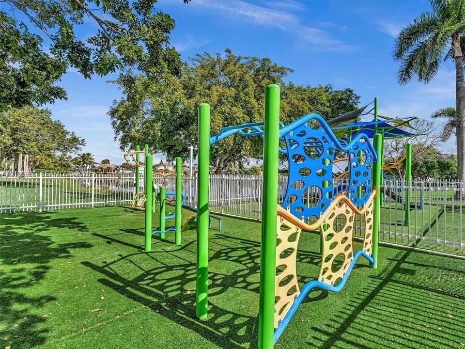 Outdoor recreation area featuring multi-element playground equipment, synthetic turf surfacing, and white perimeter fencing