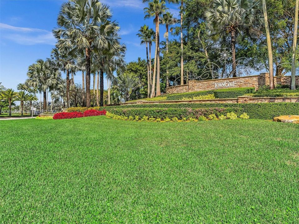 Landscaped community entrance featuring manicured lawn, tiered stone walls, mature palm trees, and ornamental metal signage