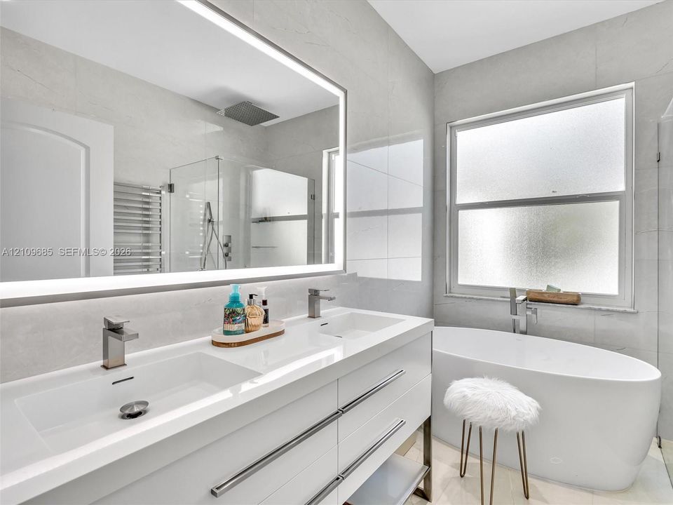 Dual-sink vanity with polished hardware, integrated LED-backlit mirror, large-format wall tiling, freestanding soaking tub, and semi-opaque window