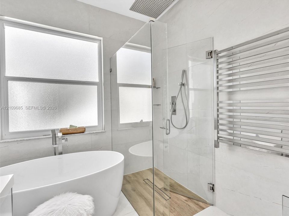 Freestanding soaking tub with chrome fixtures, walk-in glass-enclosed shower featuring a linear drain and wood-finish tile flooring, and a wall-mounted heated towel rack