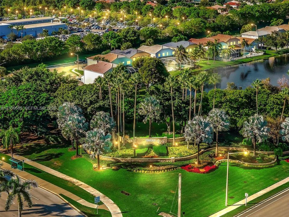 Aerial perspective of community entrance featuring stone masonry monument signage, manicured landscaping with shaped shrubbery, mature palm trees, paved pedestrian walkways, and integrated landscape lighting