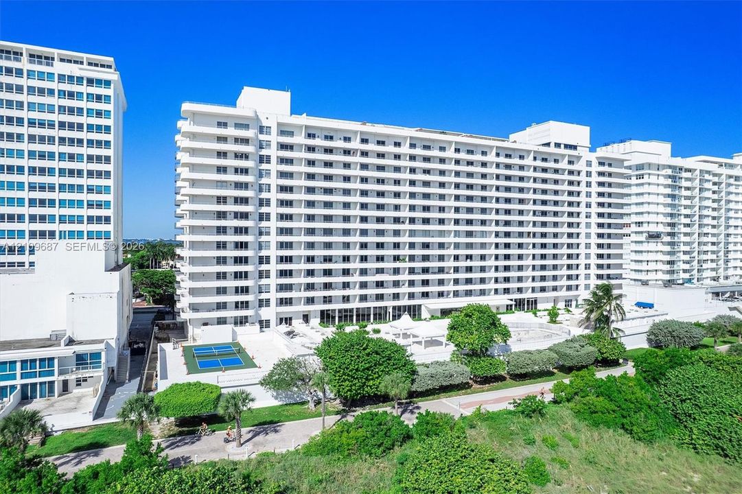 High-rise residential exterior featuring expansive tiered balconies, ground-level tennis courts, manicured landscaping, and mature palm trees