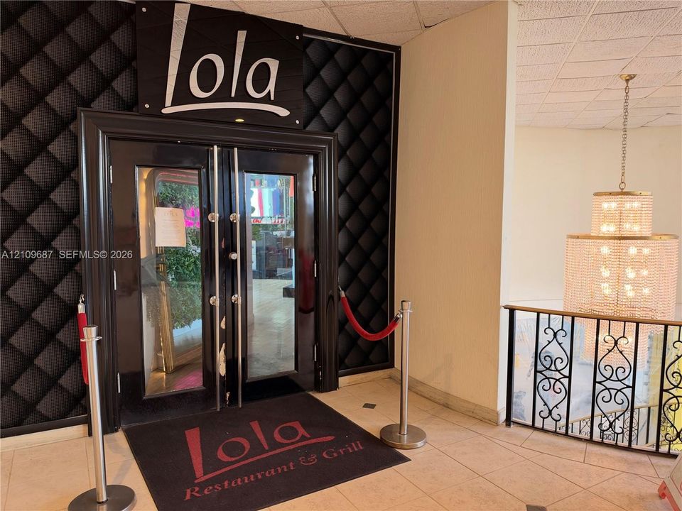 Double-door glass entrance with black finish framing, tufted black wall paneling, tile flooring, and decorative metal stair railing with integrated multi-tiered crystal chandelier