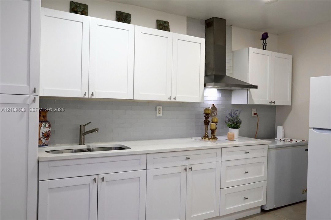 Kitchen featuring white shaker-style cabinetry, a light-toned countertop, a stainless steel chimney-style range hood, and a light gray tile backsplash