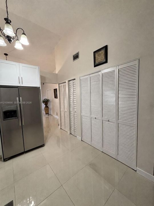 Vaulted ceiling, large-format tile flooring, stainless steel side-by-side refrigerator, white shaker-style upper cabinetry, and floor-to-ceiling louvered closet doors