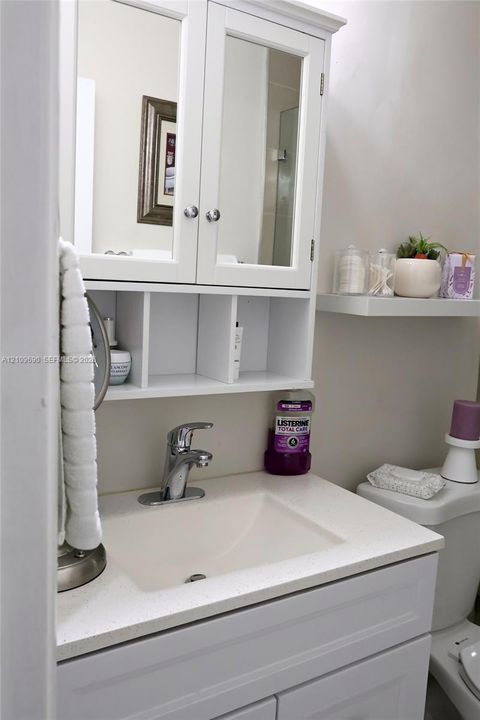 Bathroom 2 White vanity with a solid-surface countertop, integrated sink basin, chrome faucet, and mirrored cabinet featuring open display shelving