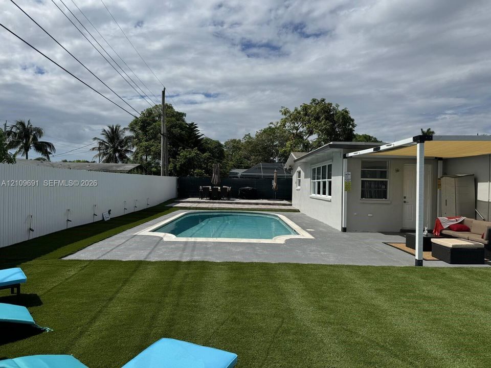 In-ground pool with concrete surround, synthetic turf lawn, white privacy fencing, and attached patio cover