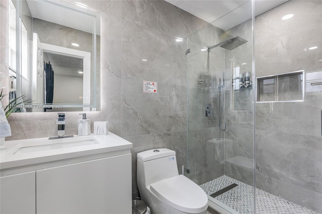 Bathroom featuring large-format gray tile wall surrounds, a frameless glass shower enclosure with a rainfall showerhead, a built-in tiled shower niche, and a white vanity with an integrated sink and backlit mirror