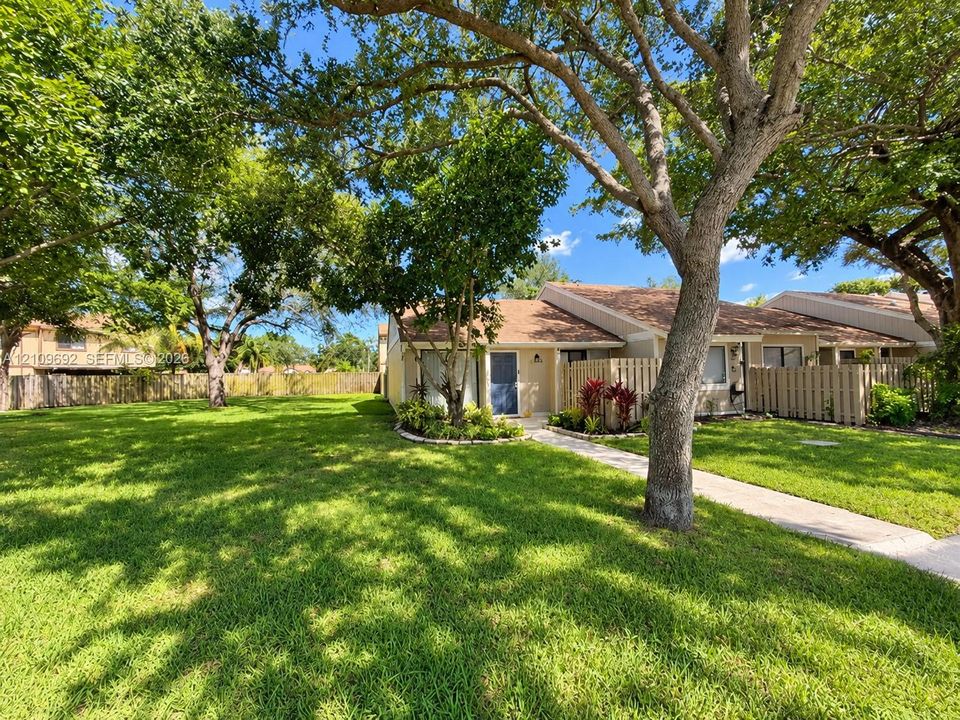 Exterior facade featuring a pitched shingle roof, concrete walkway, privacy fencing, and mature trees across a sprawling lawn