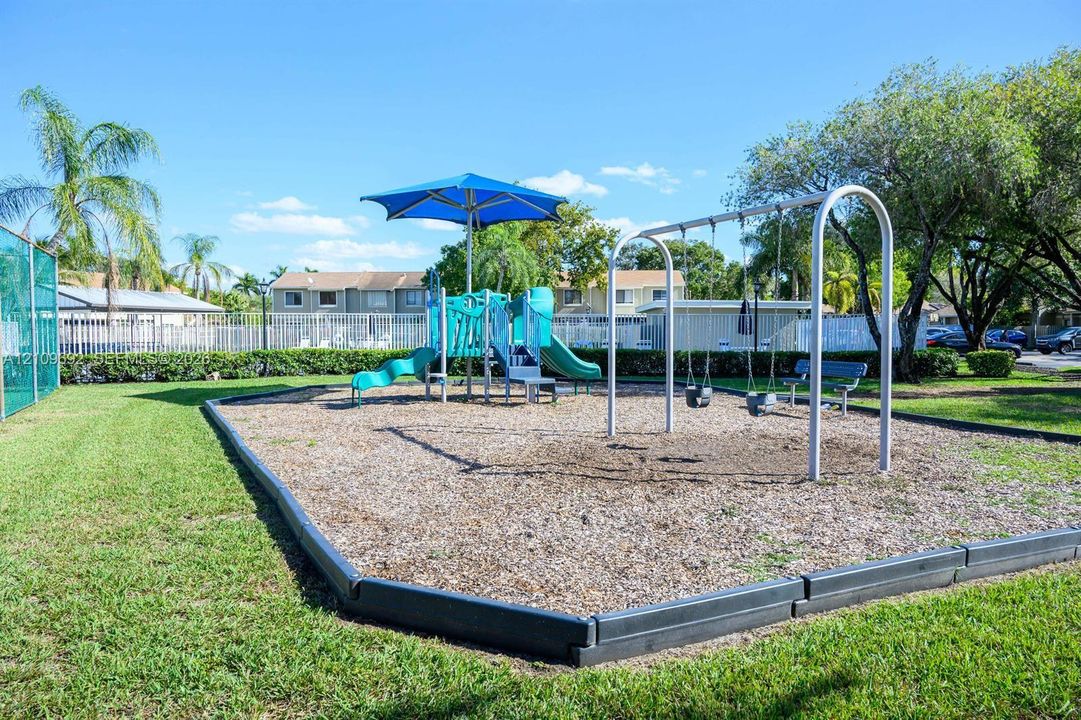 Outdoor recreation area featuring a playground structure with a blue shade canopy, two slides, a swing set, and wood-chip ground cover