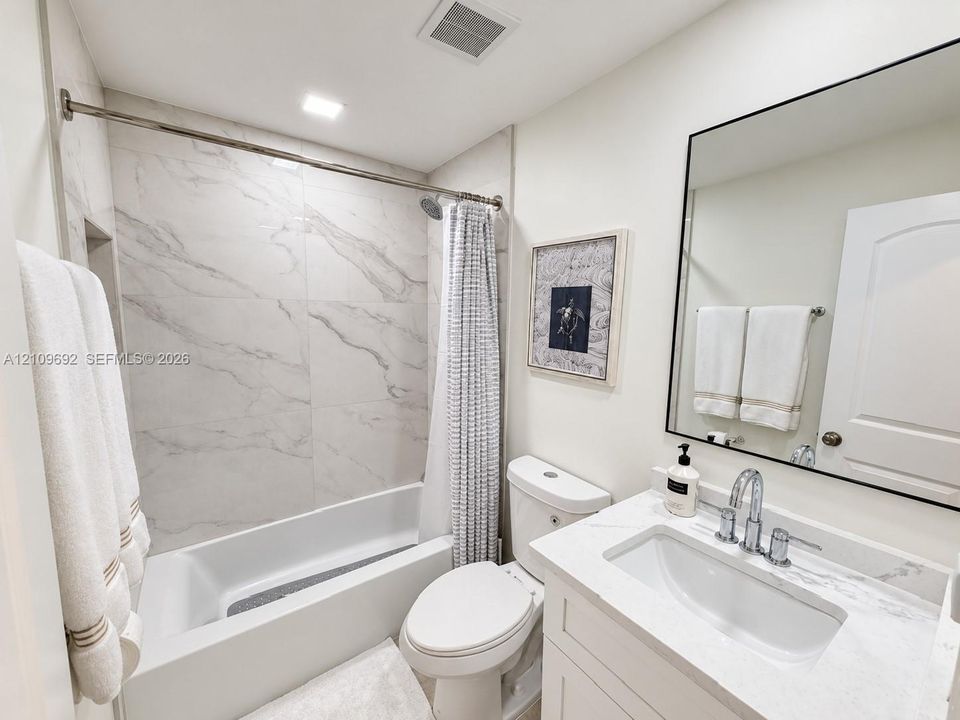 Bathroom featuring a vanity with a stone-finish countertop, undermount sink, and chrome fixtures, complemented by a large rectangular mirror with a black frame and a shower-tub combination with full-height stone-finish wall tile