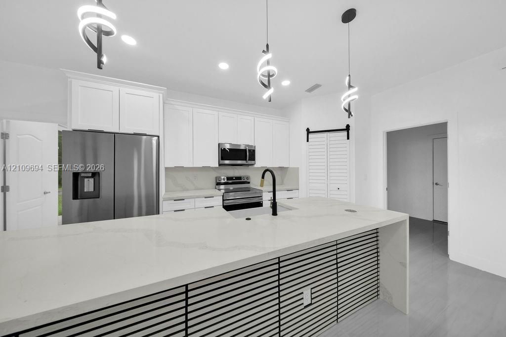 Kitchen featuring white cabinetry, an expansive island with stone-finish countertops, stainless steel appliances, matte black hardware, and contemporary spiral pendant lighting