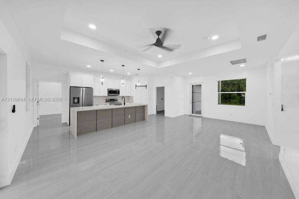 Open-concept kitchen featuring a tray ceiling, center island with horizontal slat detailing, stainless steel appliances, pendant lighting, and neutral large-format floor tiling