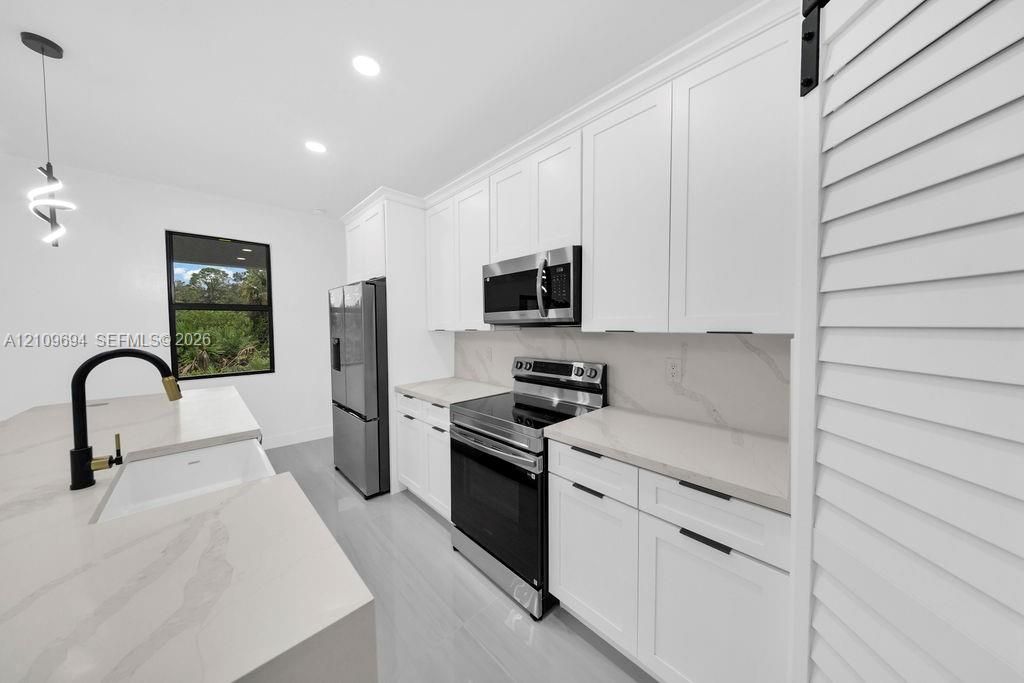 Kitchen featuring white shaker-style cabinetry, quartz countertops with integrated backsplash, stainless steel appliances, a farmhouse-style sink with a matte black faucet, and a sliding louvered barn door