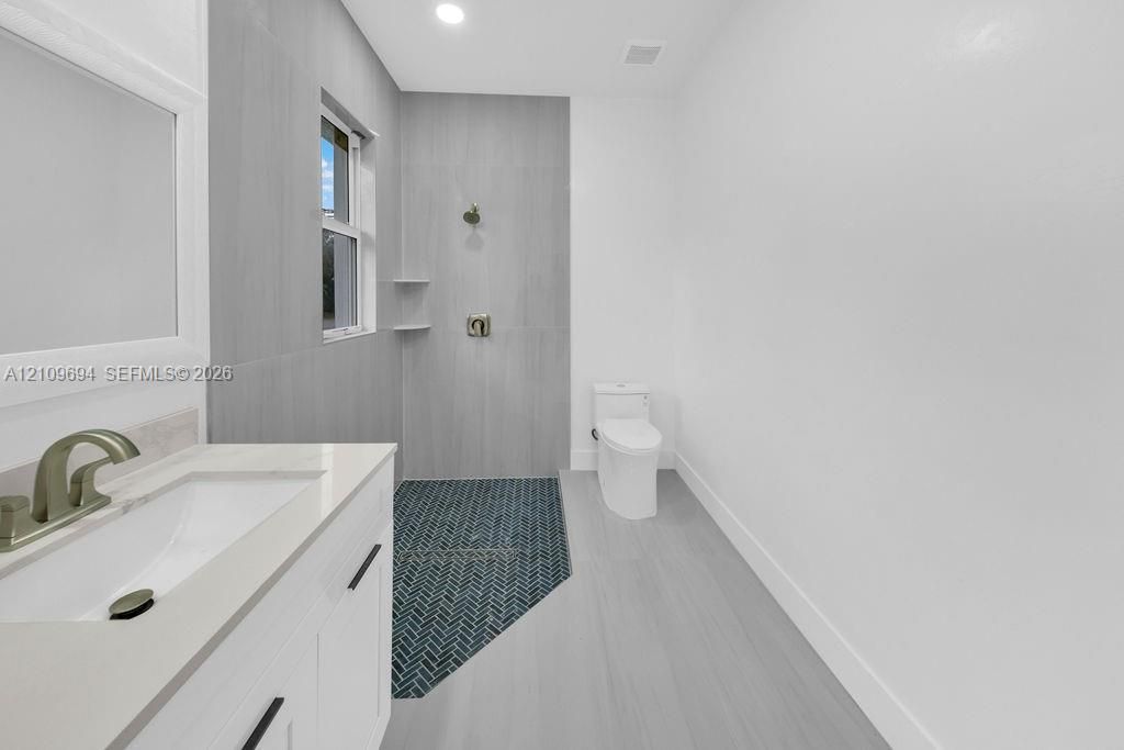 Bathroom featuring a white vanity with a stone countertop, an integrated rectangular sink, shower enclosure with floor-to-ceiling gray tile, herringbone-patterned shower floor, and a dual-flush toilet