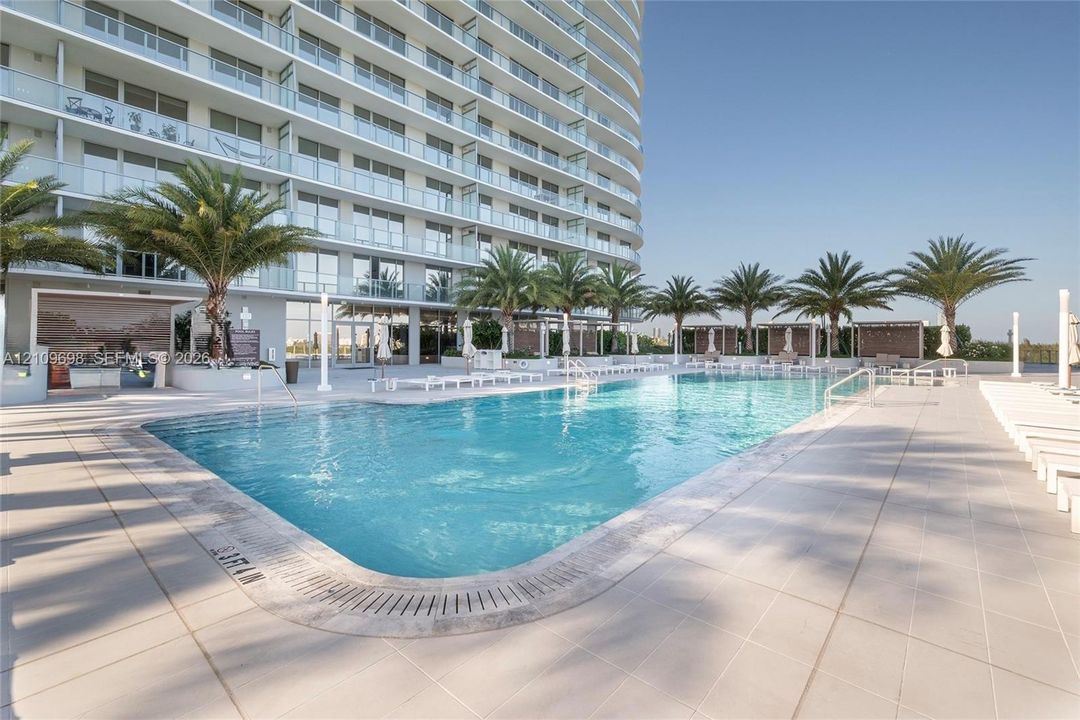 Large swimming pool with tiled surround, palm tree landscaping, and built-in outdoor kitchen area