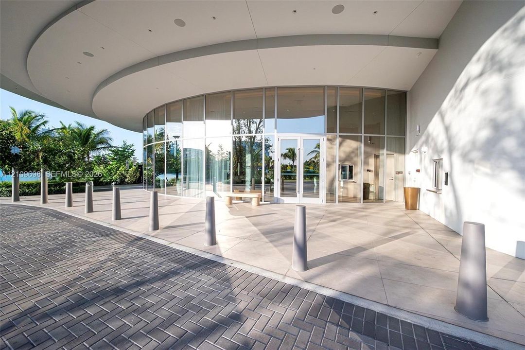 Curved glass façade featuring floor-to-ceiling windows and a deep structural overhang, complemented by a herringbone-patterned paver driveway and a wide concrete pedestrian plaza