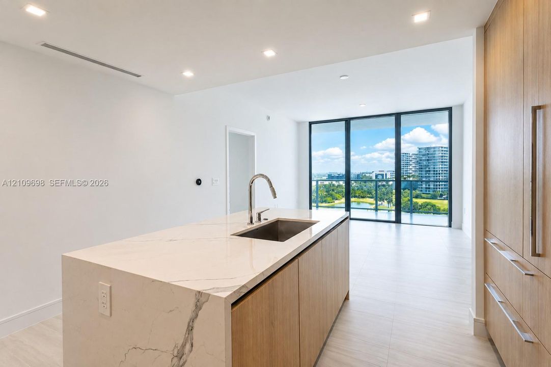 Kitchen island with stone-finish waterfall countertop, undermount stainless steel sink, wood-finish cabinetry, recessed lighting, and floor-to-ceiling glass paneling