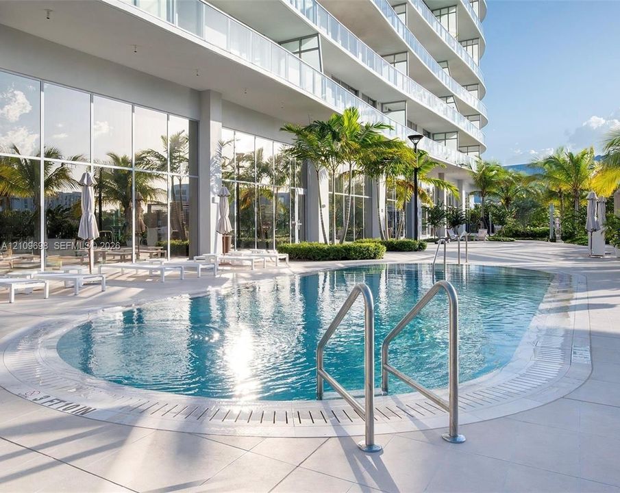 Swimming pool with metal handrails, expansive tile pool deck, floor-to-ceiling glass wall facade, and tropical landscaping