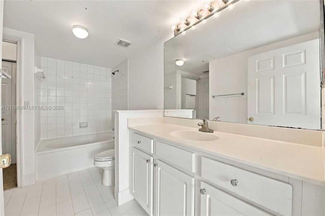 Bathroom featuring a vanity with white cabinetry, a solid surface countertop, an integrated sink, a full-width mirror, and a tiled tub surround