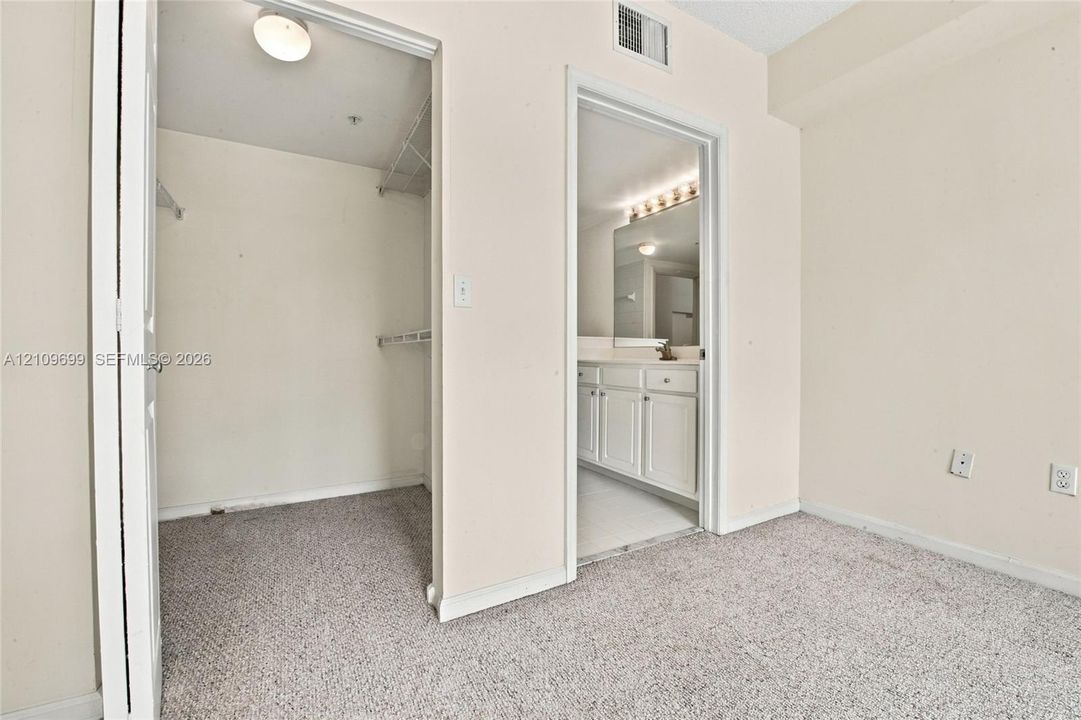 Walk-in closet featuring wire shelving and overhead lighting, with an entry to a bathroom equipped with white cabinetry, a large vanity mirror, and multi-bulb wall lighting