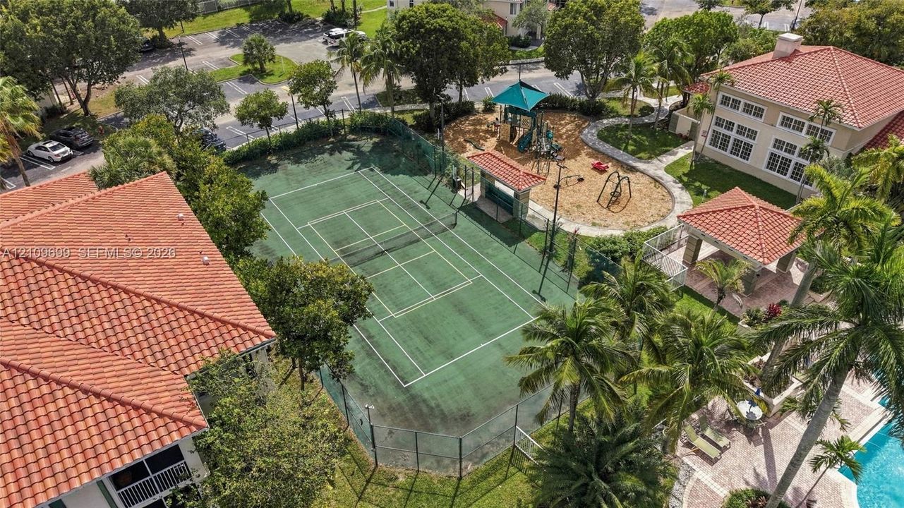 Tennis court with perimeter fencing, outdoor play area with structures, covered pavilion, and surrounding palm trees