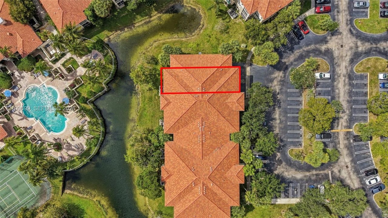 Aerial perspective of multi-unit building with a terracotta-toned shingle roof, positioned near a community swimming pool, tennis court, and water feature