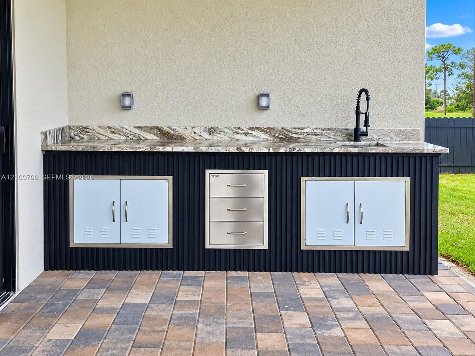Outdoor kitchen featuring a stone-look countertop, integrated stainless steel storage drawers and cabinets, a high-arc pull-down faucet, and a fluted black exterior finish on a paver patio