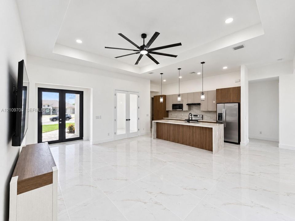 Open-concept interior featuring large-format tile flooring, a tray ceiling with a multi-blade ceiling fan, a kitchen island with a waterfall-edge countertop, two-tone cabinetry, and three integrated pendant lights