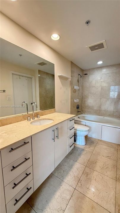 Bathroom featuring a stone countertop with an integrated sink, white cabinetry with metal hardware, a large wall-mounted mirror, a tiled bathtub surround, and stone-finish flooring
