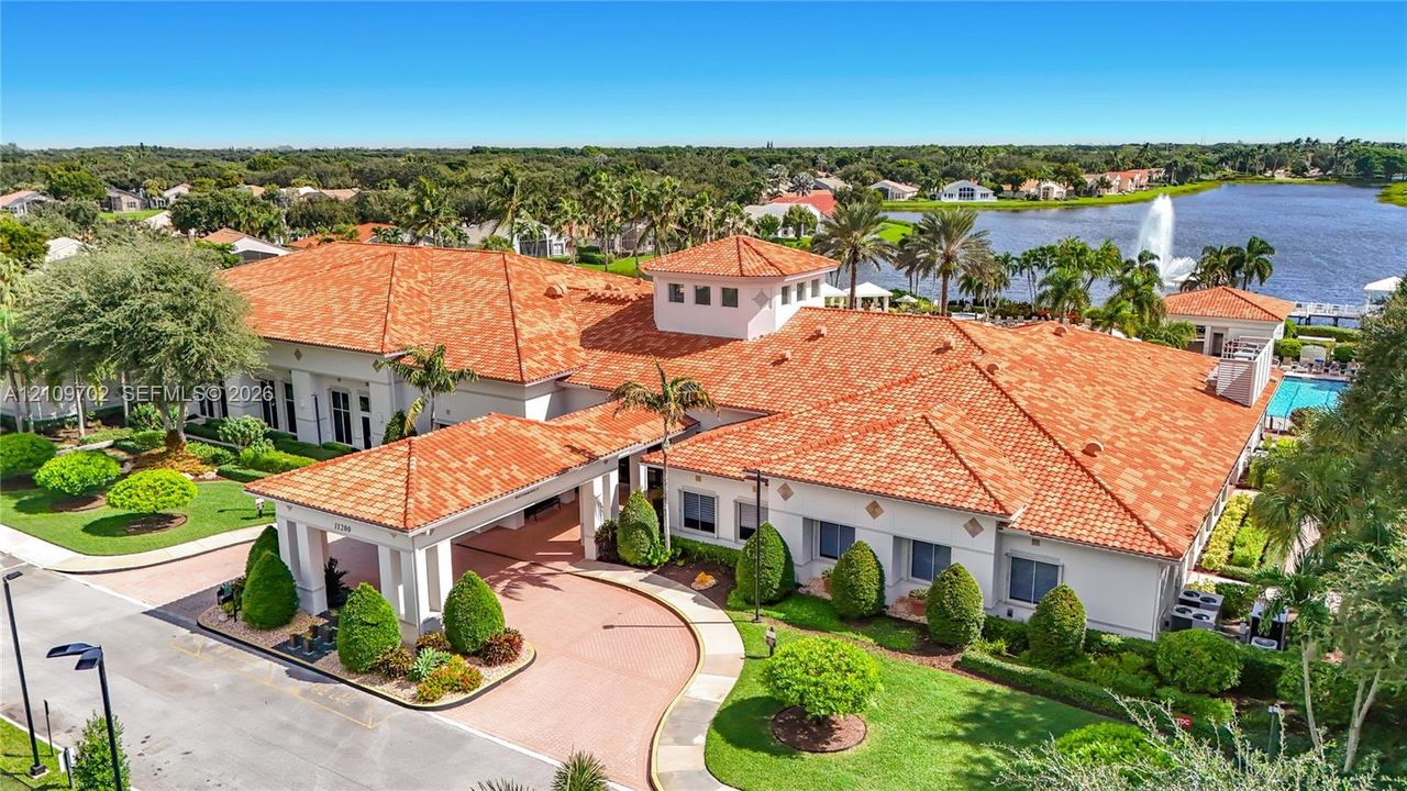 Expansive clubhouse featuring a terracotta tile roof, covered porte-cochère entry, surrounding manicured landscaping, and exterior water feature