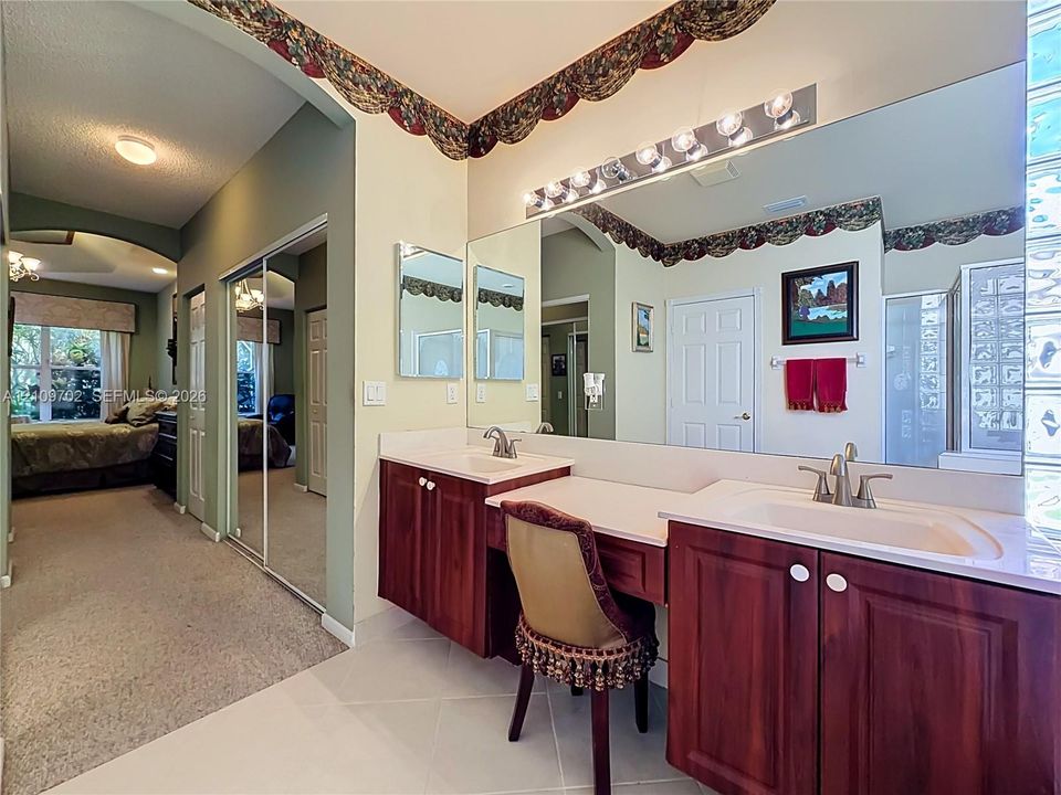 Dual vanity with wood-finish cabinetry, recessed sinks, wall-to-wall mirror, integrated seated vanity area, and glass block wall detail