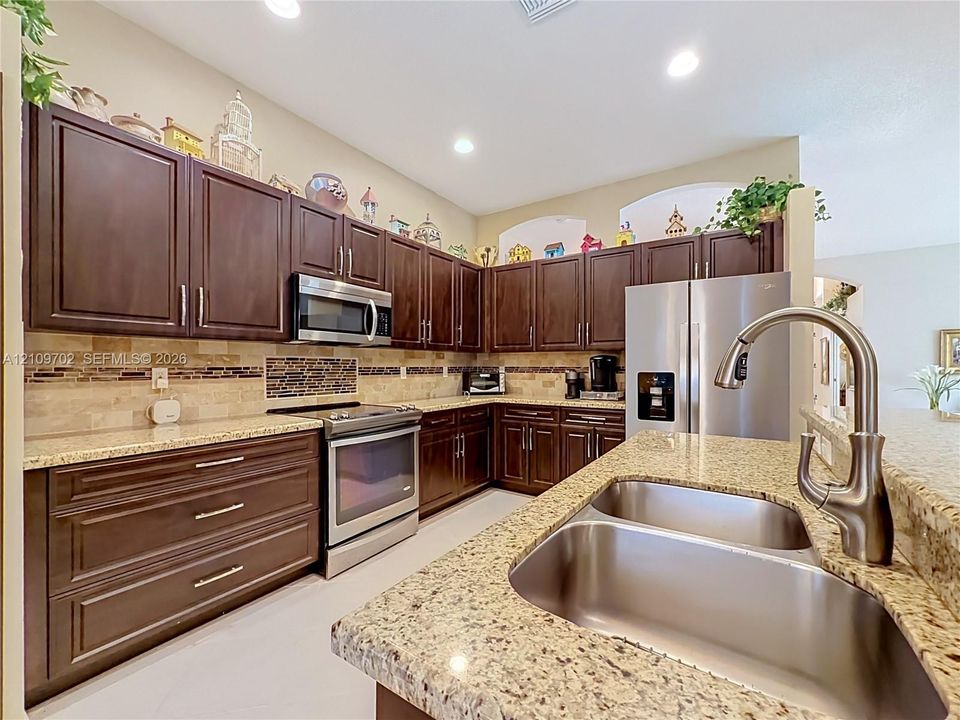 Kitchen featuring dark wood cabinetry, light stone countertops, stainless steel appliances, a double-basin undermount sink with a gooseneck faucet, and a stone tile backsplash