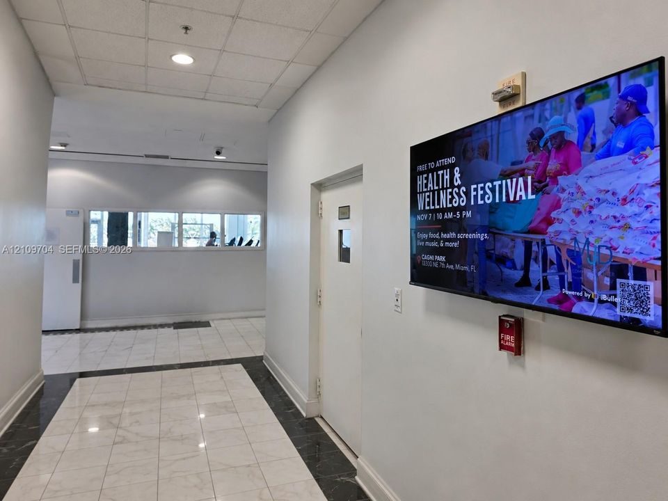Interior corridor featuring polished stone flooring with dark inlay border, recessed ceiling lighting, and wall-mounted digital display screen