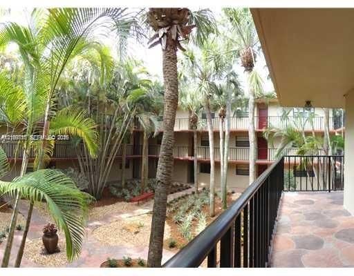Covered balcony with paver flooring and black metal railing, overlooking a courtyard featuring mature palm trees, multi-level building facade, and ground-level landscaping