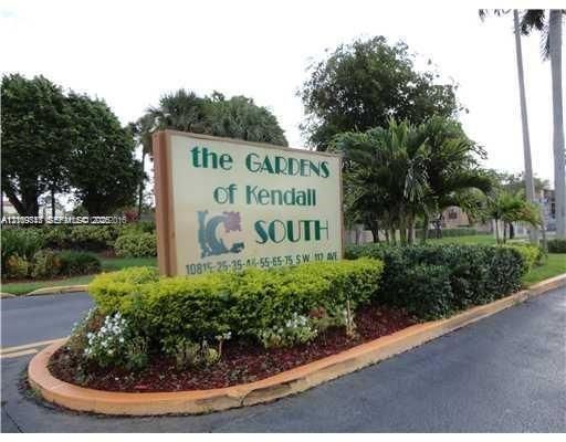 Community signage for The Gardens of Kendall South featuring manicured hedges, mature palm trees, and mulched landscaping