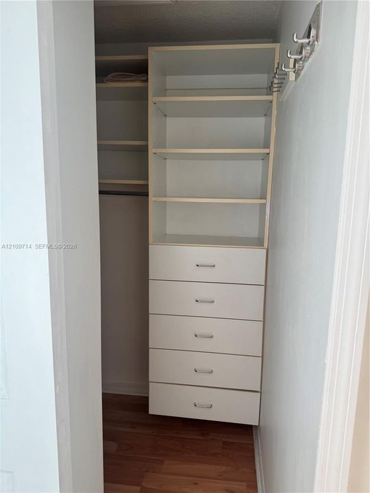 Custom reach-in closet featuring white built-in shelving, a five-drawer dresser unit with metallic pulls, a hanging rod, and wood-finish flooring