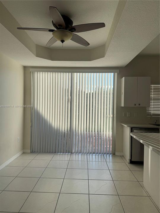 Tray ceiling with a central multi-blade ceiling fan, ceramic tile flooring, and sliding glass doors featuring vertical blinds