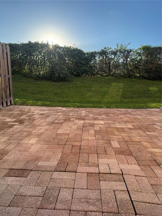 Backyard patio featuring red-toned concrete pavers, an elevated grass lawn, and a perimeter privacy hedge